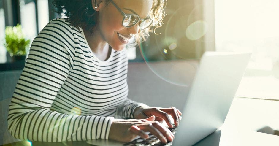 Joven mujer africana sonriente con anteojos trabajando en línea con un portátil mientras se sienta sola en una mesa