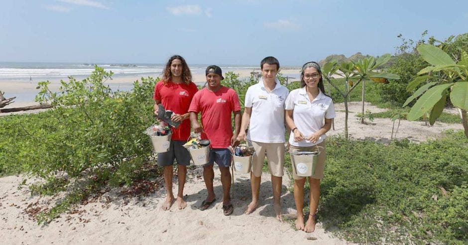 dos hombres de camisa roja, un hombre da camisa blanca y una mujer de camisa blanca posan con baldes de aluminio en una playa