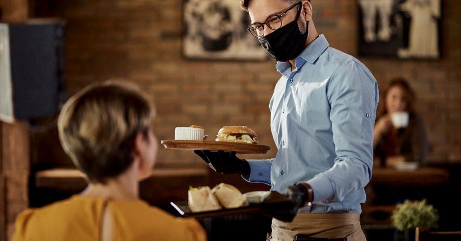 un hombre con mascarilla sirve el almuerzo a un comensal en un restaurante