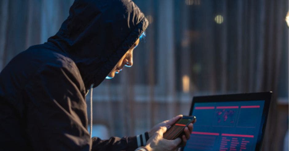 hombre con sudadera con capucha sosteniendo celular frente a una laptop