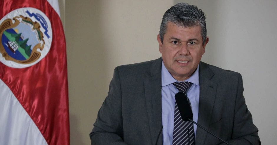 un hombre de saco y corbata habla frente a un micrófono. Hay una bandera de costa rica al fondo.