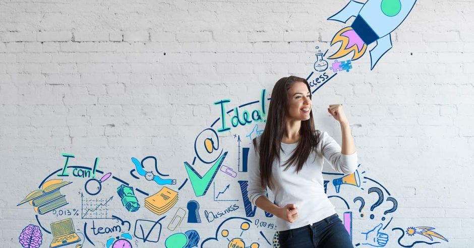 mujer joven con puño cerrado y brazo encogido, detrás de ella pared con dibujos referentes a ideas de negocio
