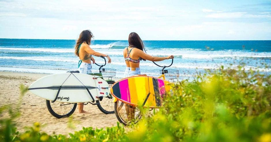 dos mujeres en bicicleta llevan consigo una tabla de surf