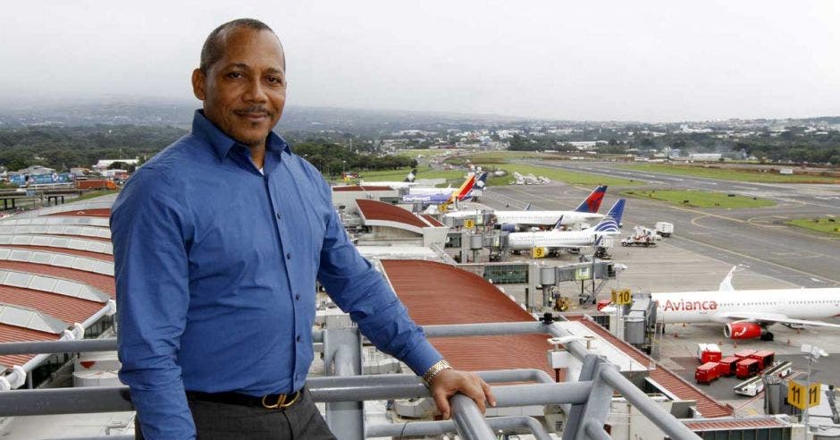 un hombre de camisa azul y pantalón gris posa sobre el fondo de un aeropuerto en operaciones