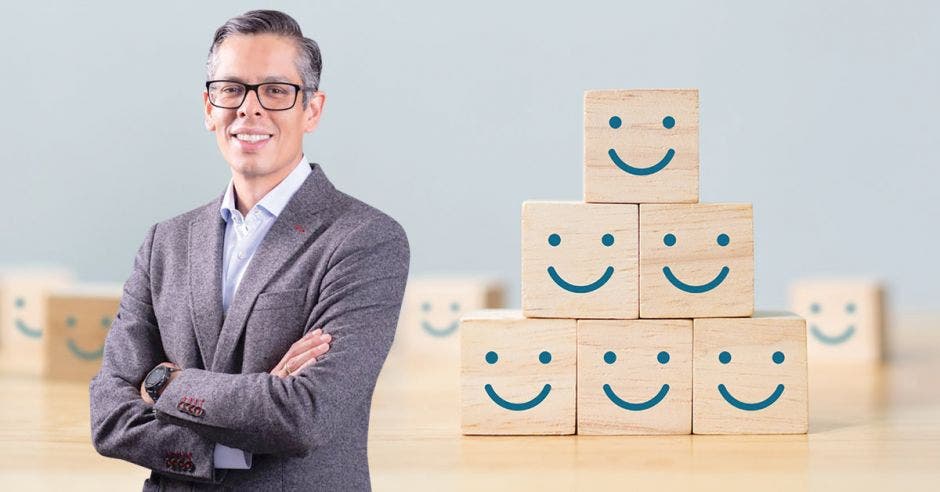 hombre de lentes con traje gris, cruzado de brazos, de fondo cubos de madera con caritas felices