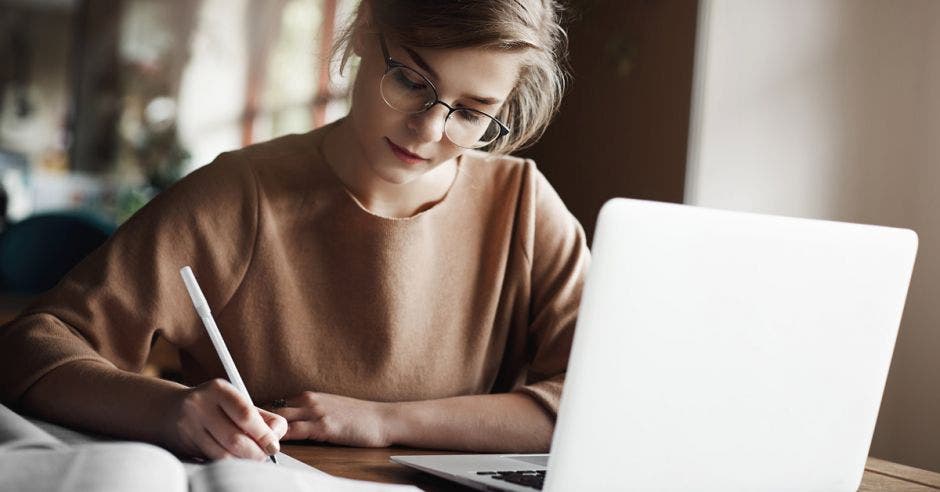 Mujer con blusa café escribiendo con lapicero y al frente una computadora blanca