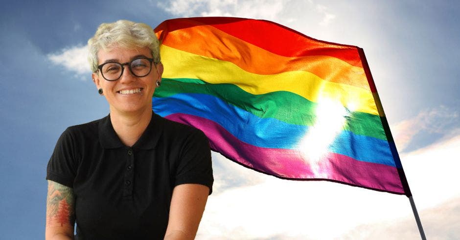 Una mujer con blusa y lentes negros, pelo blanco posando y sonriendo. Atrás la bandera de la diversidad.